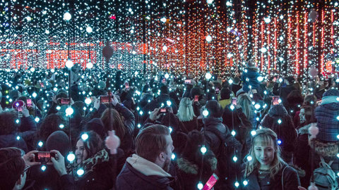 A large group of people stand under neon lights suspended by wire. Each wire has approximately 10 blue globes on it. People are crowded underneath them smiling and taking photos